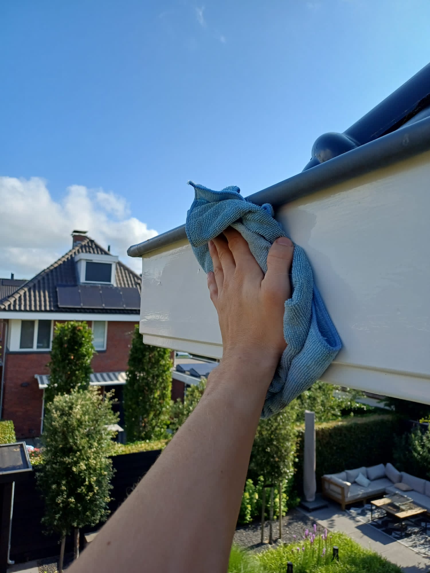 Hand met een blauwe doek die een witte daklijst schoonmaakt, met achtertuinen op de achtergrond